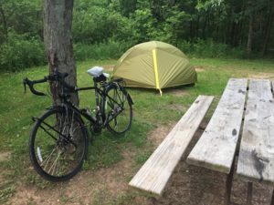 Foreground: an empty picnic table, a black touring bike with a plastic bag around the saddle leaning on a tree. Behind them is a green tent with bushes behind that.