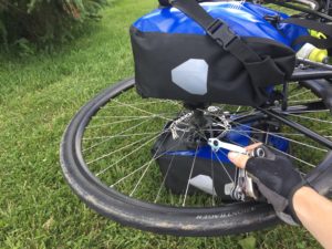 The front wheel of a bike with panniers still attached to the rack. A hand is holding up a spoke tool in front of the wheel.